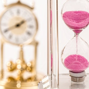 Puede incluir: Un reloj de arena transparente con arena rosa que fluye de la bombilla superior a la inferior. El reloj de arena se encuentra sobre un fondo borroso con un reloj dorado.