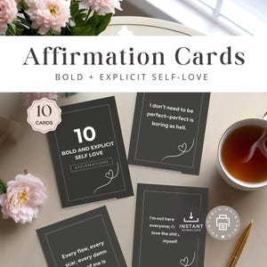 May include: A set of 10 affirmation cards with the text "Affirmation Cards" and "Bold + Explicit Self-Love". The cards are dark gray with white text and feature various affirmations. A cup of tea and flowers are in the background.