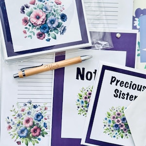 May include: A collection of stationery items featuring floral designs. Includes notecards with a purple border, a pen engraved with "Mindy", and notebooks. The notecards have a floral bouquet design with pink, blue, and purple flowers.