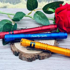 May include: A collection of four colorful flashlights in red, blue, and gold, each with a black pocket clip. The blue flashlight has the text "Crafted By Cynthia". The flashlights are arranged on a wooden slice with a red rose and green leaves in the background.