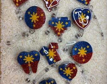May include: A collection of glittery badge reels in various shapes, including hearts, circles, and a tooth. The designs feature the colors of the Philippine flag: blue, red, and yellow. Each reel has a clear plastic retractable cord.