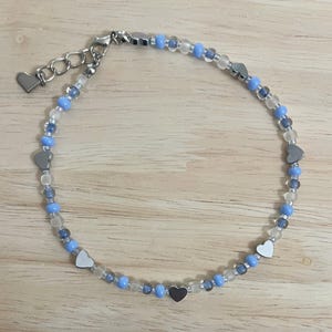 Könnte beinhalten: Ein zartes Fußkettchen mit einer silbernen Kette und blauen und klaren Perlen. Die Perlen sind mit kleinen silbernen Herzanhängern durchsetzt.