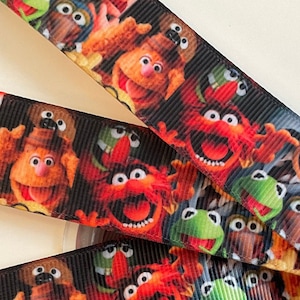 May include: Black ribbon featuring a colorful pattern of Muppet characters, including Kermit the Frog and Animal. The ribbon is wrapped around a white spool, with several strands visible.