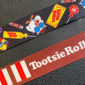 Op de afbeelding: Twee rollen lint, een zwart met een witte uil en kleurrijke snoepontwerpen, de andere bruin met witte strepen en de tekst "Tootsie Roll" in wit.