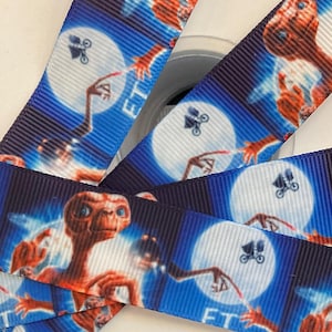 May include: A roll of blue ribbon featuring the character E.T. against a starry night sky background. The ribbon also includes the text "E.T." and a graphic of a bicycle with a basket. The ribbon is wrapped around a white spool.