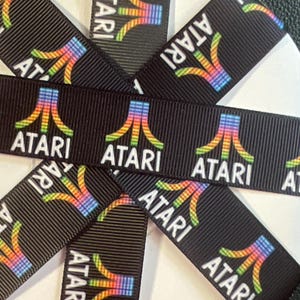 Pode incluir: Uma fita de gorgorão preta enrolada em torno de um carretel circular branco. A fita apresenta um padrão repetitivo do logótipo Atari num gradiente arco-íris, com a palavra Atari impressa em letras brancas sob cada logótipo.