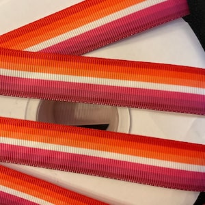 May include: A close-up of three rolls of lesbian pride ribbon. The ribbon is a multi-colored striped pattern of pink, orange, white, and red.