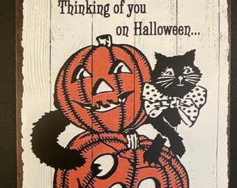 May include: A vintage-style metal sign with a white background and black and orange illustrations. The sign features two pumpkins with smiling faces and a black cat wearing a white bow tie. The text on the sign reads "Thinking of you on Halloween..."