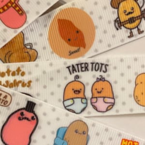 Puede incluir: Cinta blanca con un patrón repetido de patatas de dibujos animados en varias poses y atuendos. Las patatas llevan sombreros, sostienen pistolas y están sentadas en sofás. El texto "Tater Tots", "Harry Potato", "Fry Baby", "Potato Jalrus", "I'm a Potato", "Couch Potato" y "Hot Potato!" está impreso en la cinta.