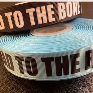 May include: Two rolls of ribbon with the text "Bad to the Bone" printed on them. One roll is black with white text and the other is light blue with black text.