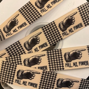 May include: Tan ribbon with a black and white houndstooth pattern. The ribbon features a repeating design of a pointing hand with the text "PULL MY FINGER!" in black. The ribbon is spooled on a white roll.