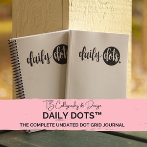 Puede incluir: Dos cuadernos blancos con espiral y letras negras que dicen "daily dots" en la portada. Los cuadernos están sobre una superficie de madera. El texto "TB Calligraphy & Design DAILY DOTS™ THE COMPLETE UNDATED DOT GRID JOURNAL" se muestra sobre un fondo rosa.