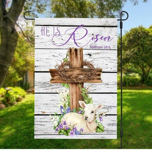May include: A white wooden garden flag with a brown wooden cross, a wreath of thorns, and a white lamb lying at the foot of the cross. The flag has the text "He is Risen" in purple lettering and "Matthew 28:6" in black lettering.