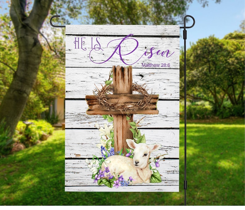 May include: A white wooden garden flag with a brown wooden cross, a wreath of thorns, and a white lamb lying at the foot of the cross. The flag has the text "He is Risen" in purple lettering and "Matthew 28:6" in black lettering.