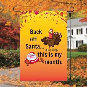 May include: A yellow and orange garden flag with a cartoon turkey and Santa Claus. The text on the flag reads "Back off Santa... this is my month."