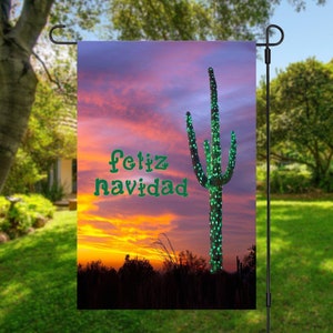 May include: A garden flag with a sunset background and a tall cactus decorated with green lights. The text "Feliz Navidad" is written in green on the flag.