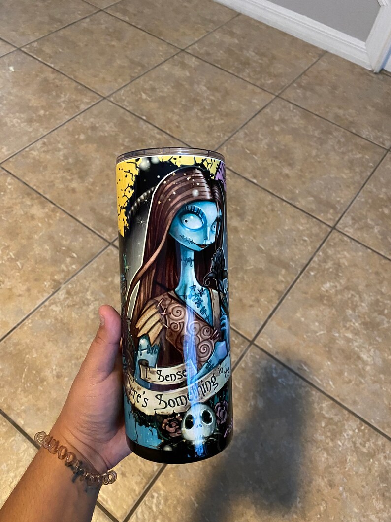 May include: A black and blue stainless steel tumbler featuring a graphic of Sally from The Nightmare Before Christmas. The tumbler has a white and gold rim and the text "There's Something About You" in a banner.