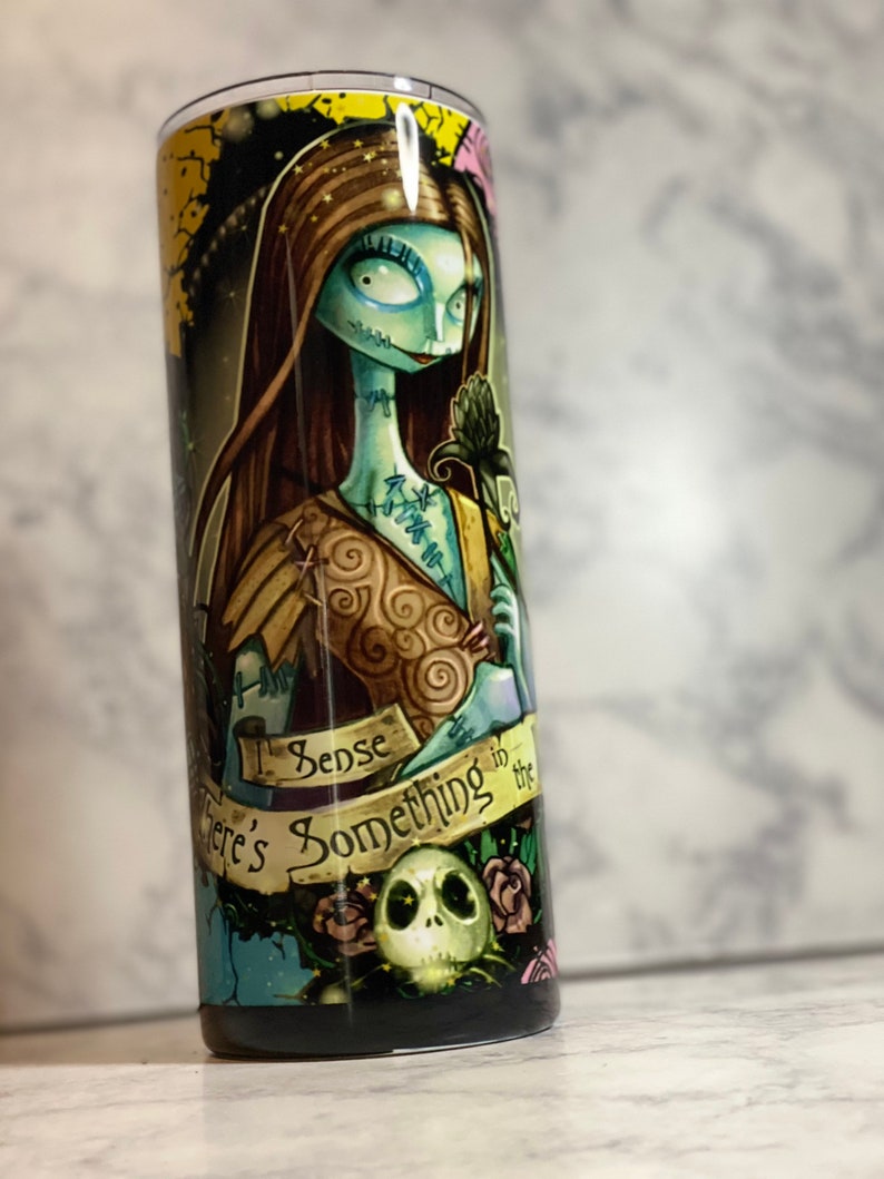 May include: A black and green stainless steel tumbler featuring a graphic of Sally from The Nightmare Before Christmas. The tumbler has the text "I sense something in the air" on it.