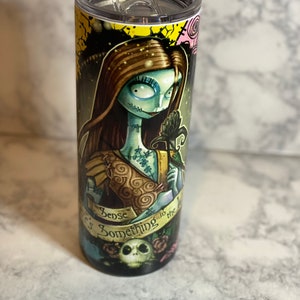 May include: A stainless steel tumbler featuring a colourful graphic of a character from the film "The Nightmare Before Christmas". The graphic depicts a woman with long brown hair, a blue dress, and a black rose. The text "There's Something in the Woods" is printed on the tumbler.
