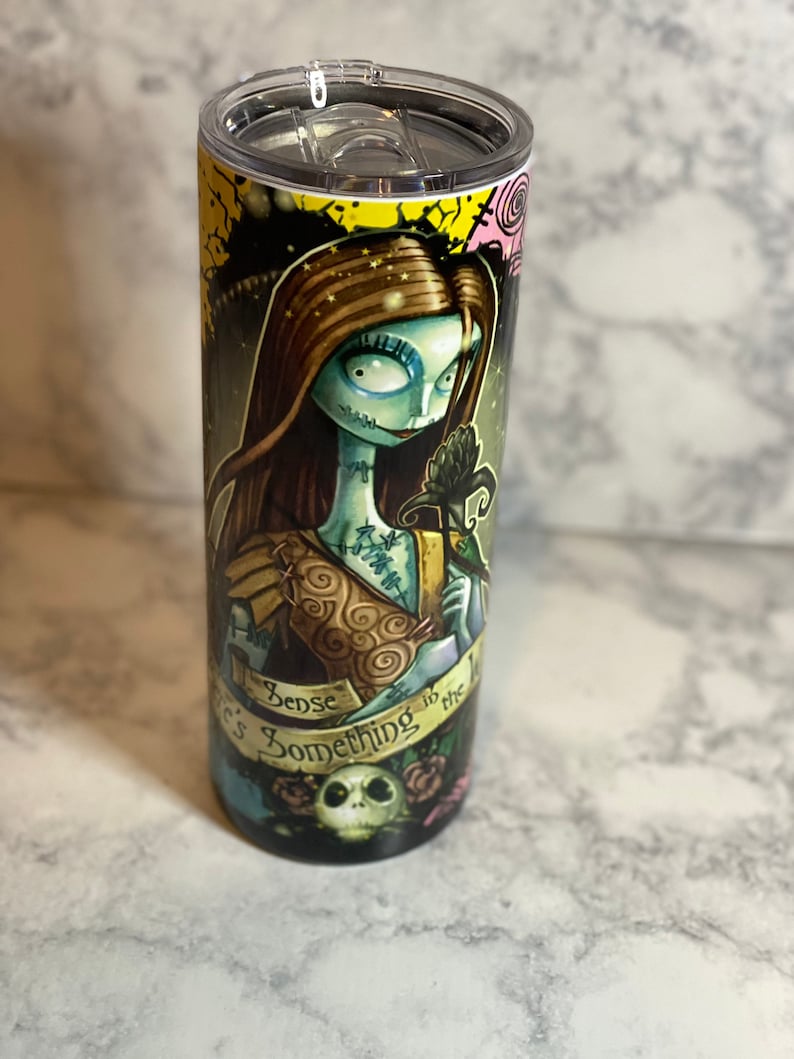 May include: A stainless steel tumbler featuring a colourful graphic of a character from the film "The Nightmare Before Christmas". The graphic depicts a woman with long brown hair, a blue dress, and a black rose. The text "There's Something in the Woods" is printed on the tumbler.