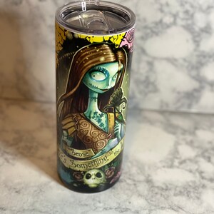May include: A stainless steel tumbler featuring a colourful graphic of a character from the film "The Nightmare Before Christmas". The graphic depicts a woman with green skin and brown hair, holding a flower. The text "There's Something in the Air" is printed on the tumbler.