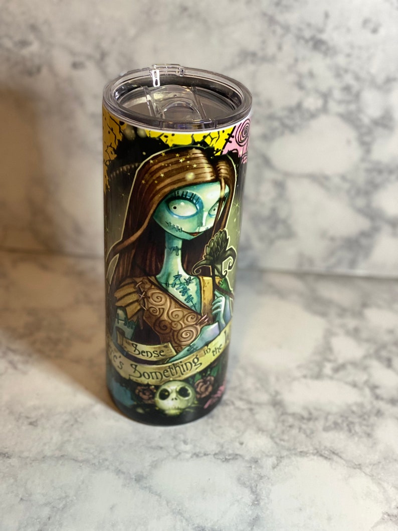 May include: A stainless steel tumbler featuring a colourful graphic of a character from the film "The Nightmare Before Christmas". The graphic depicts a woman with green skin and brown hair, holding a flower. The text "There's Something in the Air" is printed on the tumbler.