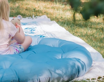 May include: A large, round, tufted blue floor cushion placed on a white and blue patterned blanket. The cushion features a soft, padded surface with visible circular stitching details, providing a comfortable seating area for relaxation.