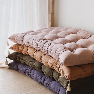 May include: Four floor cushions stacked on top of each other. The cushions are made of a soft, textured fabric and have a tufted design. The cushions are in shades of pink, brown, green, and purple. Each cushion has a white tassel on the corner.
