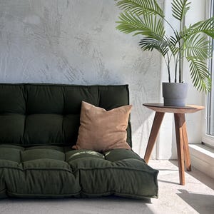 Pode incluir: Um futon de veludo cotelê verde escuro com uma almofada castanho-claro. Uma pequena mesa lateral de madeira suporta uma planta verde em vaso. O fundo é uma parede cinza texturizada e uma janela.