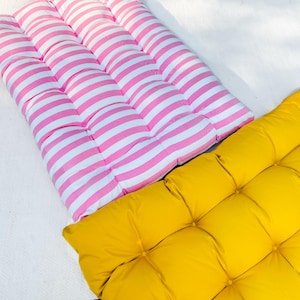Puede incluir: Dos cojines de suelo, uno de rayas rosa y blanco y otro de color amarillo s&oacute;lido. Ambos cojines est&aacute;n acolchados y tienen forma cuadrada.