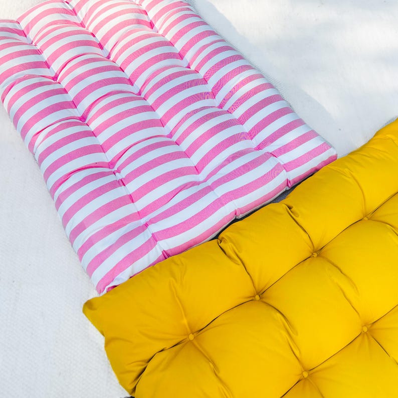 Puede incluir: Dos cojines de suelo, uno de rayas rosa y blanco y otro de color amarillo s&oacute;lido. Ambos cojines est&aacute;n acolchados y tienen forma cuadrada.