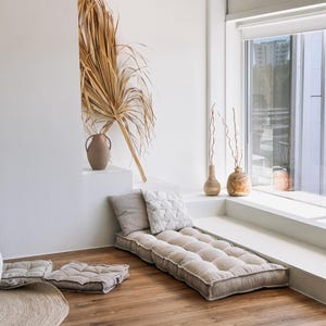 May include: A neutral-toned floor cushion and pillows are arranged on a wooden floor. A long, rectangular cushion is paired with two square cushions and two decorative pillows. A dried palm frond and vases add to the room's decor.