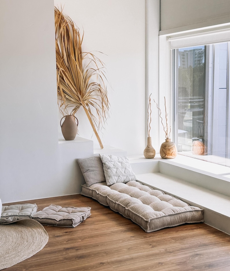 May include: A neutral-toned floor cushion and pillows are arranged on a wooden floor. A long, rectangular cushion is paired with two square cushions and two decorative pillows. A dried palm frond and vases add to the room's decor.