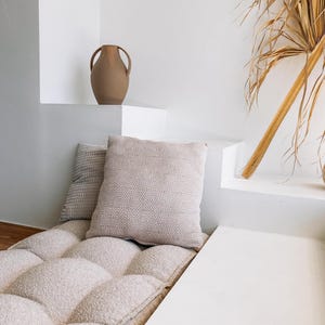 May include: A neutral-toned, tufted cushion with two decorative pillows and a brown vase. The pillows have textured and geometric patterns. The vase has two handles. A dried palm frond leans against the white wall.