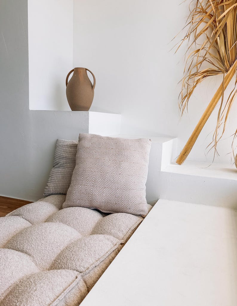 May include: A neutral-toned, tufted cushion with two decorative pillows and a brown vase. The pillows have textured and geometric patterns. The vase has two handles. A dried palm frond leans against the white wall.