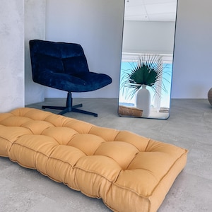May include: An interior shot featuring a mustard-colored, tufted floor cushion. A navy blue swivel chair is positioned to the left, and a tall, arched mirror reflects a vase with greenery. The room has a minimalist aesthetic.