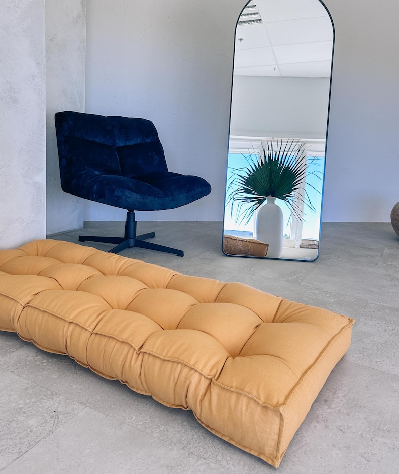 May include: An interior shot featuring a mustard-colored, tufted floor cushion. A navy blue swivel chair is positioned to the left, and a tall, arched mirror reflects a vase with greenery. The room has a minimalist aesthetic.