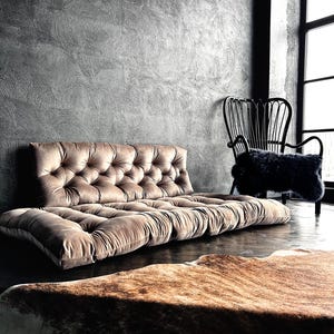 May include: A tufted, low-profile sofa in a muted brown hue dominates the foreground. The sofa sits on a dark wooden floor, with a cowhide rug in front. A black chair with a furry seat and a decorative chair are in the background.