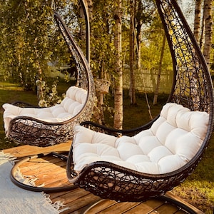 Könnte beinhalten: Zwei braune Hängesessel aus Weidengeflecht mit weißen Kissen auf einem Holzdeck. Die Sessel hängen an einem Metallgestell und sind von Bäumen umgeben.
