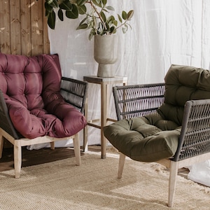 May include: Two outdoor armchairs with woven backs and light wood legs. One chair has a burgundy cushion, the other has an olive green cushion. A small side table with a concrete vase is between the chairs. water repellent