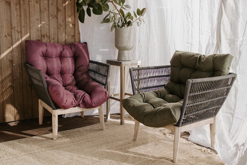 May include: Two outdoor armchairs with woven backs and light wood legs. One chair has a burgundy cushion, the other has an olive green cushion. A small side table with a concrete vase is between the chairs. water repellent