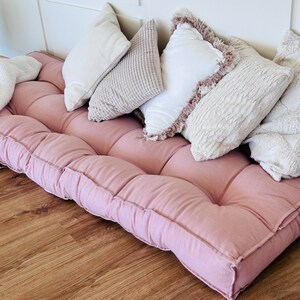 Puede incluir: Un coj&iacute;n de suelo rosa con m&uacute;ltiples secciones acolchadas y una variedad de almohadas decorativas en tonos blancos y beige.
