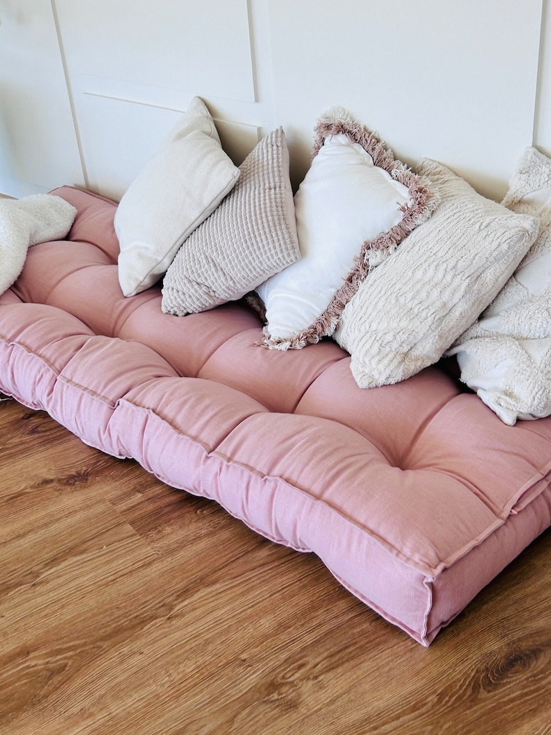Puede incluir: Un coj&iacute;n de suelo rosa con m&uacute;ltiples secciones acolchadas y una variedad de almohadas decorativas en tonos blancos y beige.