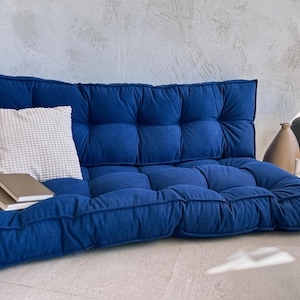 May include: A deep blue, tufted floor cushion with a matching backrest. A white textured pillow and two books rest on the cushion. Decorative vases are in the background.