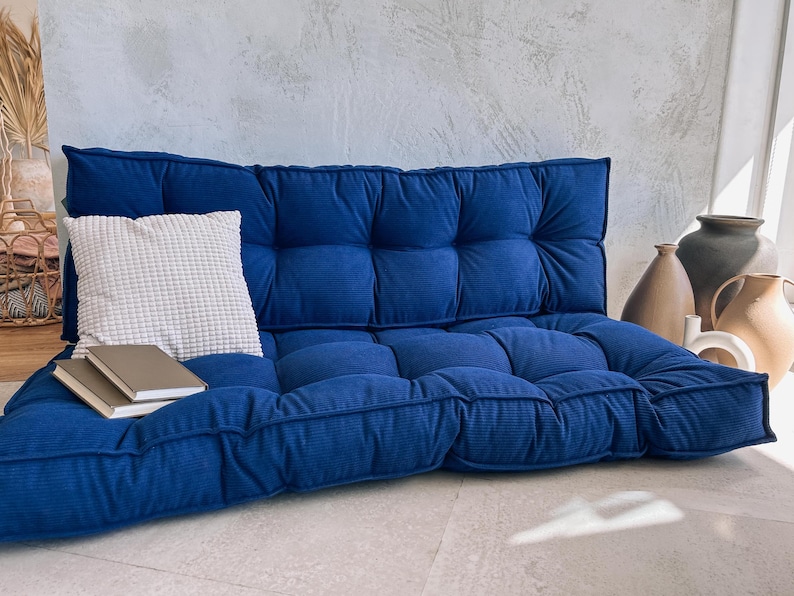 May include: A deep blue, tufted floor cushion with a matching backrest. A white textured pillow and two books rest on the cushion. Decorative vases are in the background.