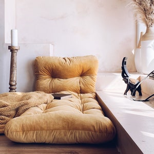 May include: A mustard yellow floor cushion with a tufted top sits on a wooden floor in front of a white wall. A fluffy brown throw blanket is draped over the cushion. Two wooden candle holders with white candles are on the floor to the left of the cushion.