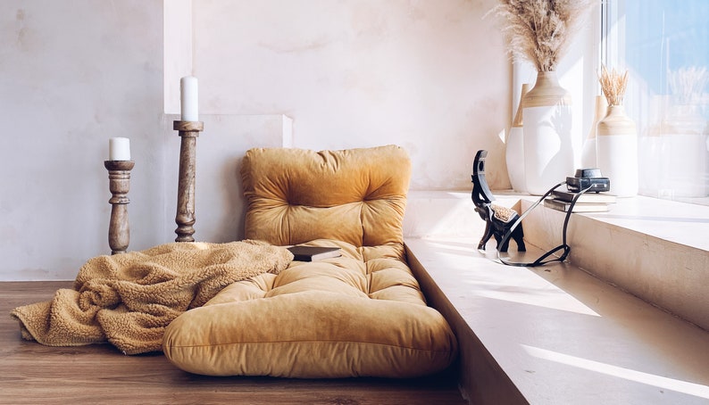 May include: A mustard yellow floor cushion with a tufted top sits on a wooden floor in front of a white wall. A fluffy brown throw blanket is draped over the cushion. Two wooden candle holders with white candles are on the floor to the left of the cushion.