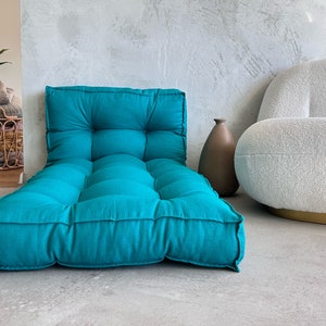 Puede incluir: Un coj&iacute;n de suelo azul turquesa con botones capiton&eacute;s y forma rectangular. El coj&iacute;n est&aacute; sobre un suelo de hormig&oacute;n gris claro.