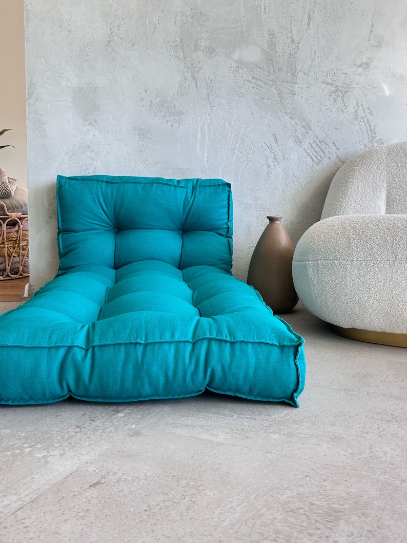 Puede incluir: Un coj&iacute;n de suelo azul turquesa con botones capiton&eacute;s y forma rectangular. El coj&iacute;n est&aacute; sobre un suelo de hormig&oacute;n gris claro.
