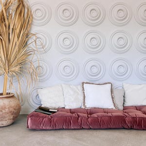 May include: A living room scene featuring a low, burgundy velvet sofa with several white and beige pillows. A large, dried palm frond is in a terracotta pot. The wall has a repeating circular pattern.